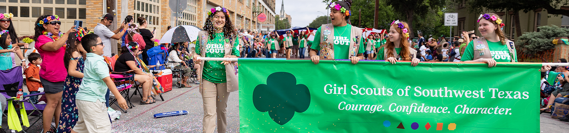  Girl Scouts smiling and holding a green banner in the battle of flowers parade while a boy throws confetti on them 