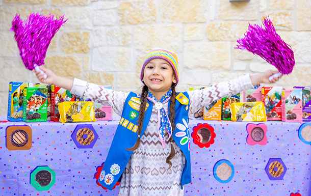 girl scout jumps with pom-poms in hand in front of a cookie booth